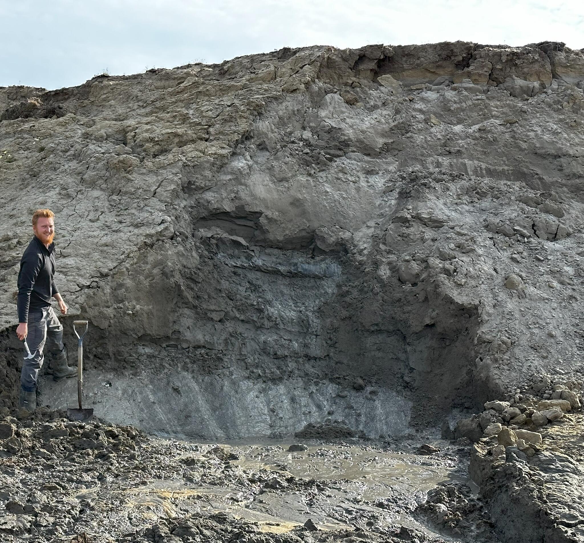 Sam stands in front of a slab of gray rock, wearing a black shirt and gray pants, holding a hammer in one hand and a shovel in the other.
