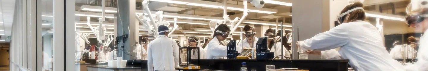 Group of students in a laboratory room, all wearing white lab gear.
