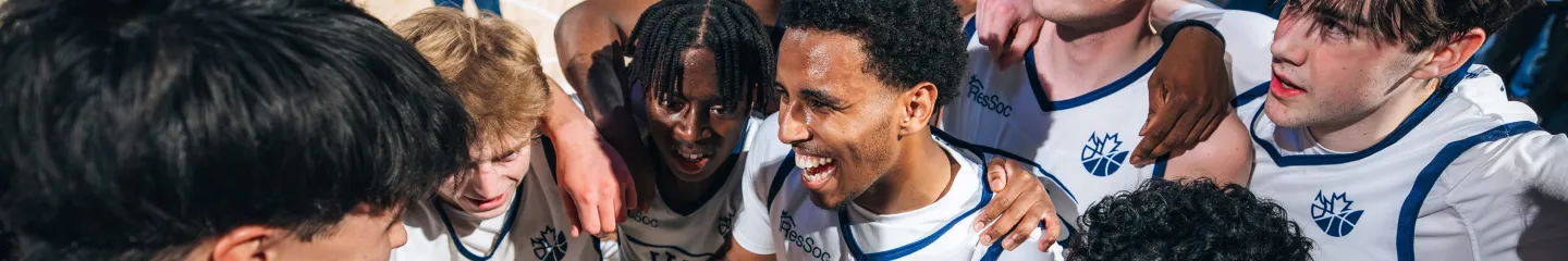 Photo of a group of university athletes in white Queen’s ResSoc basketball jerseys with blue trim huddle closely together on an indoor court, with arms around each other’s shoulders.