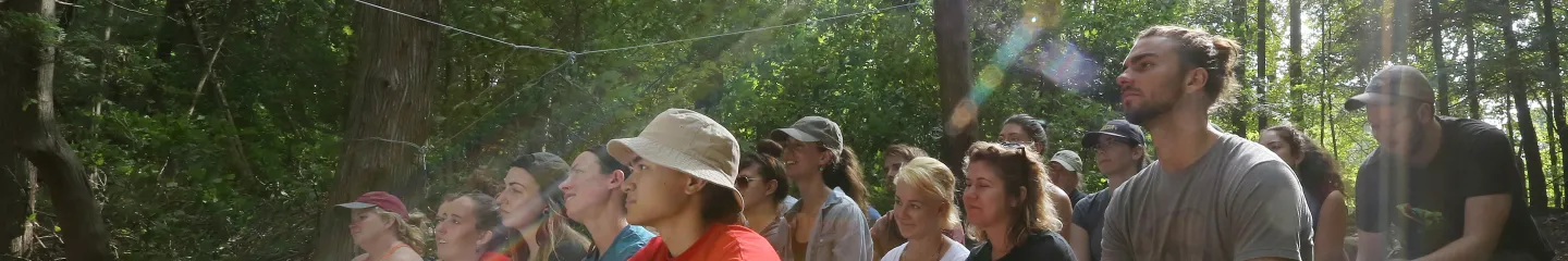 A diverse group of individuals seated on the forest floor, engaged in a discussion about outdoor education and experiential learning.