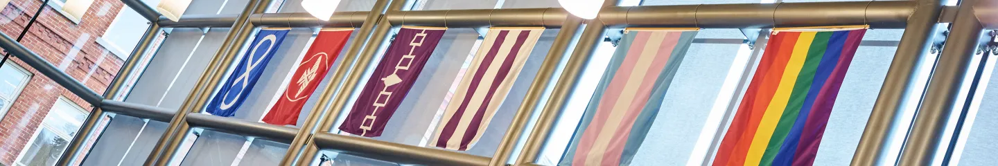 Six flags – Anishinaabe First Nations People, Six Nations Confederacy, Two Row Wampum, Métis Nation, LGBTQ2S+ Pride, and Trans Pride hang in windows of Goodes Hall