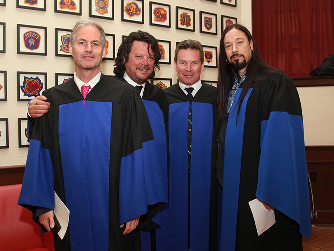 Four members of the Tragically Hip stand together in Grant Hall wearing blue-and-black honorary degree gowns. They face the camera and smile in front of a wall displaying framed Queen's University crests. One member holds a white program, while another rests an arm around a bandmate's shoulder. 