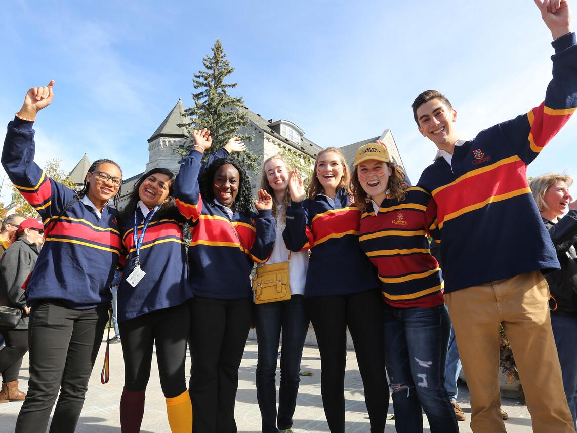Seven Queen’s University students in striped rugby shirts cheer with raised arms at a sunny campus celebration in front of a stone building.
