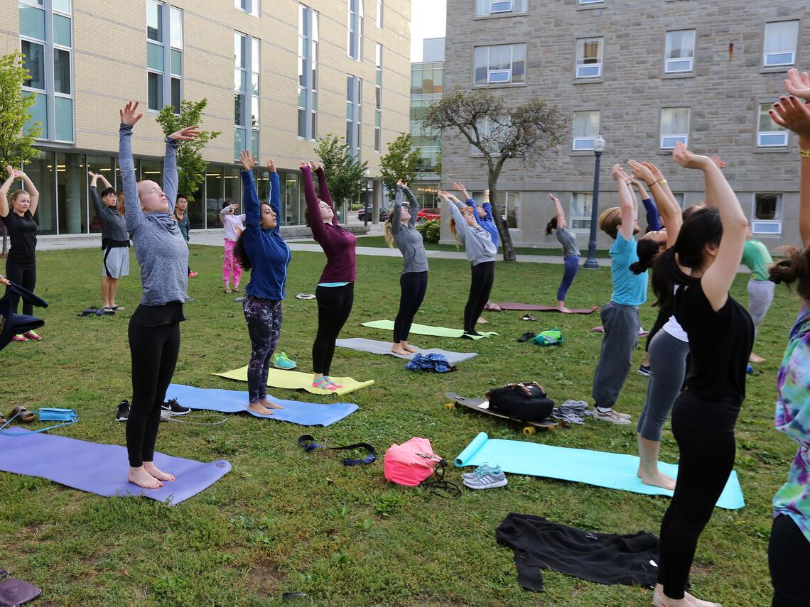 Students participate in an outdoor group yoga class on a grassy lawn between campus buildings, standing on mats with their arms raised overhead.