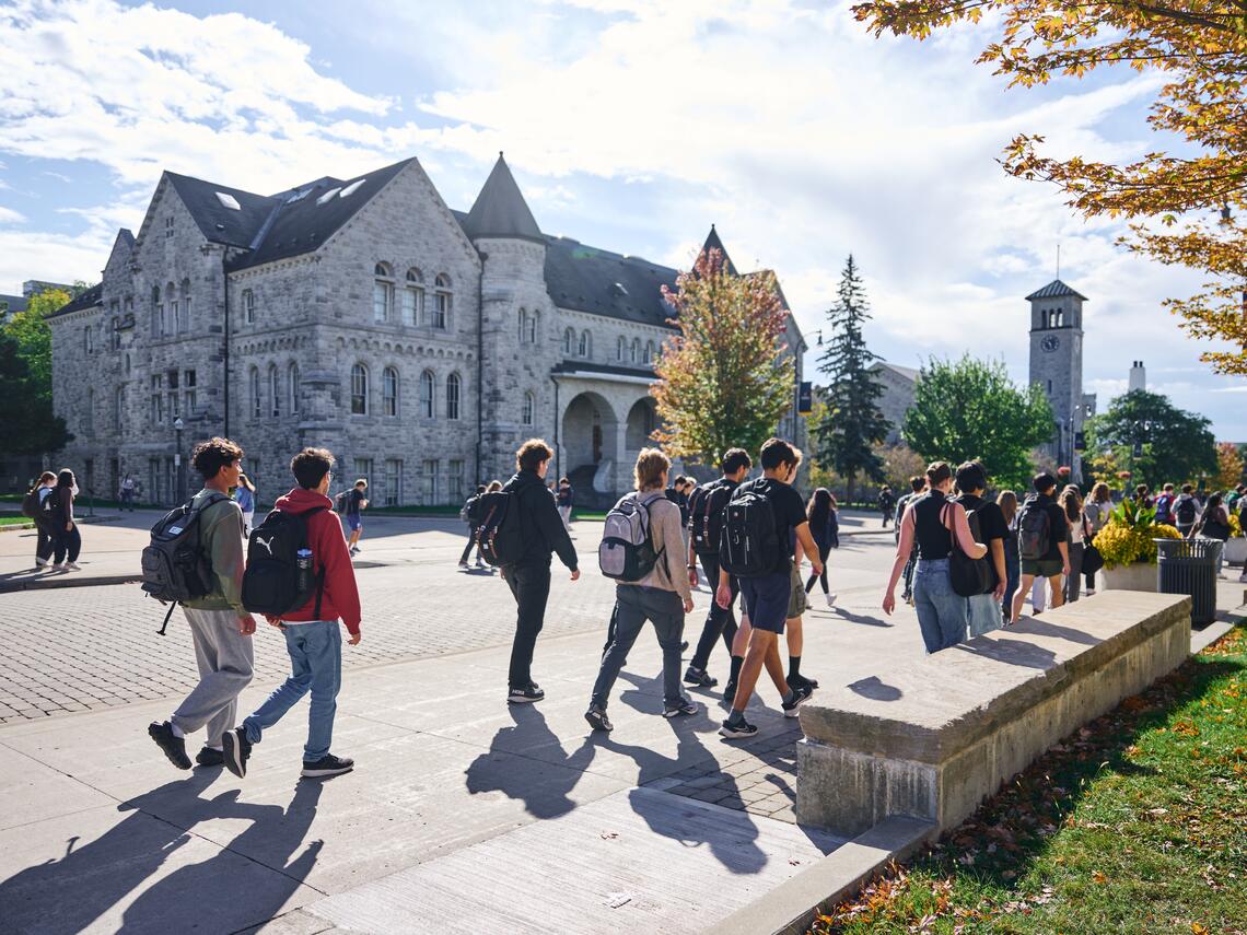 Students with backpacks walk in front of Ontario Hall at Queen's university surrounded by trees with early autumn leaves.