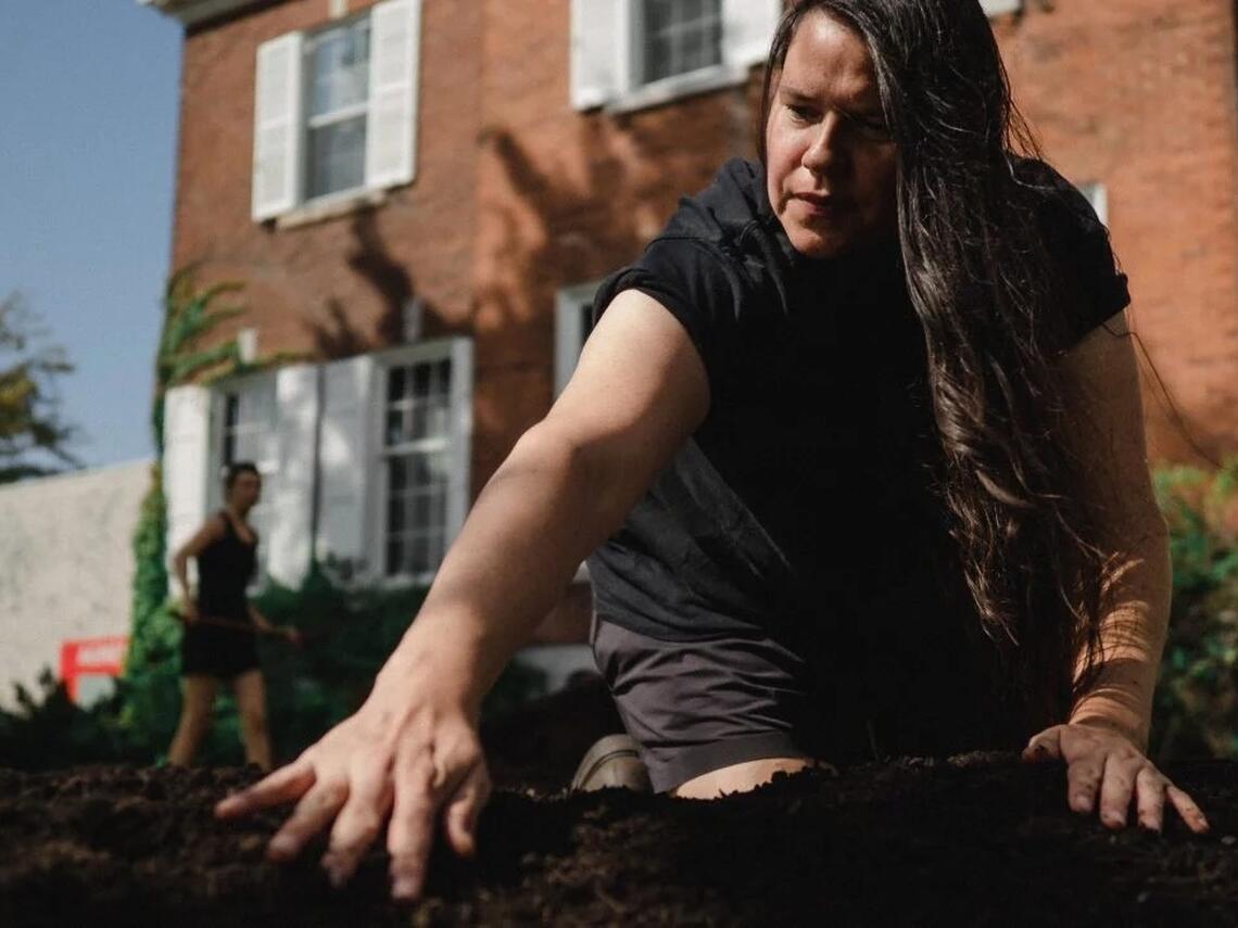 Artist Anne Riley kneels in dark soil outside the Agnes Etherington Art Centre at Queen’s University, reaching across a garden bed while installing Dark Matter Garden.   https://agnes.queensu.ca/connect/news-and-stories/anne-riley-installs-dark-matter-garden/