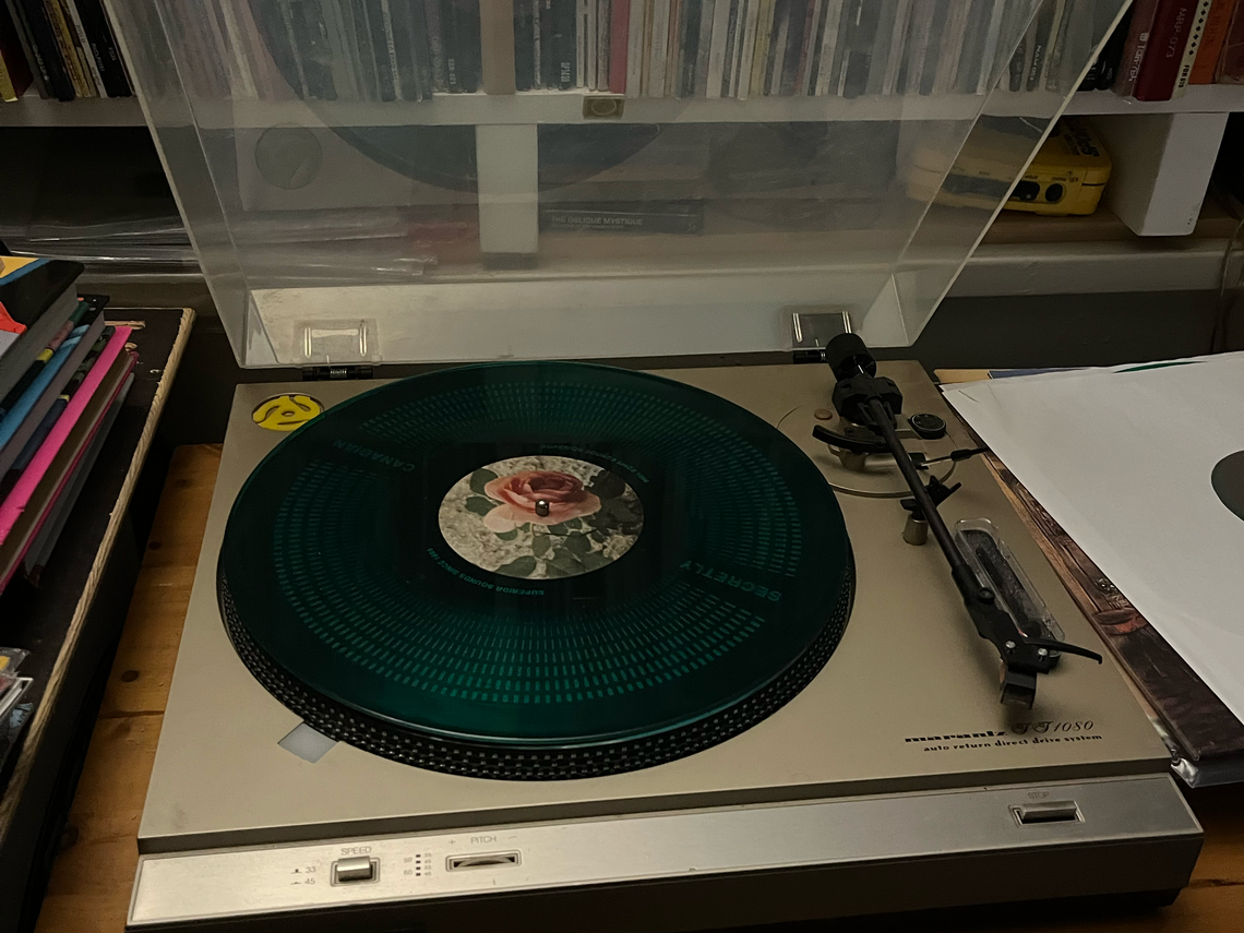 A record player holds the record, Angel Olson Big Time, a dark green transparent record with a vintage looking pink rose in the centre
