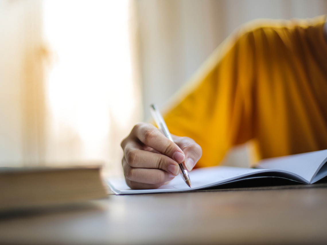 Close-up of a person writing with a pen in an open notebook at a desk. A stack of books is blurred in the foreground, and the person's face is out of frame.
