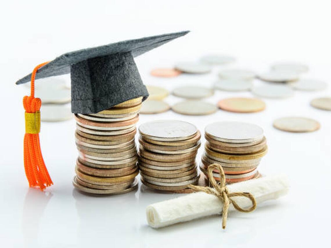 Stacked coins arranged in ascending height with a graduation cap on the tallest stack and a rolled diploma tied with string in the foreground, symbolizing the financial cost or investment of higher education.