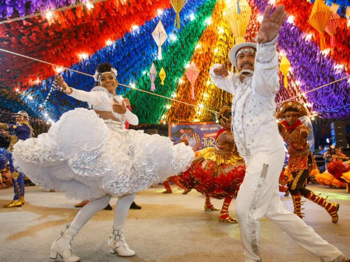 Dancers in colorful costumes perform under a rainbow canopy of paper decorations and lights at a festive Brazilian June festival celebration. 