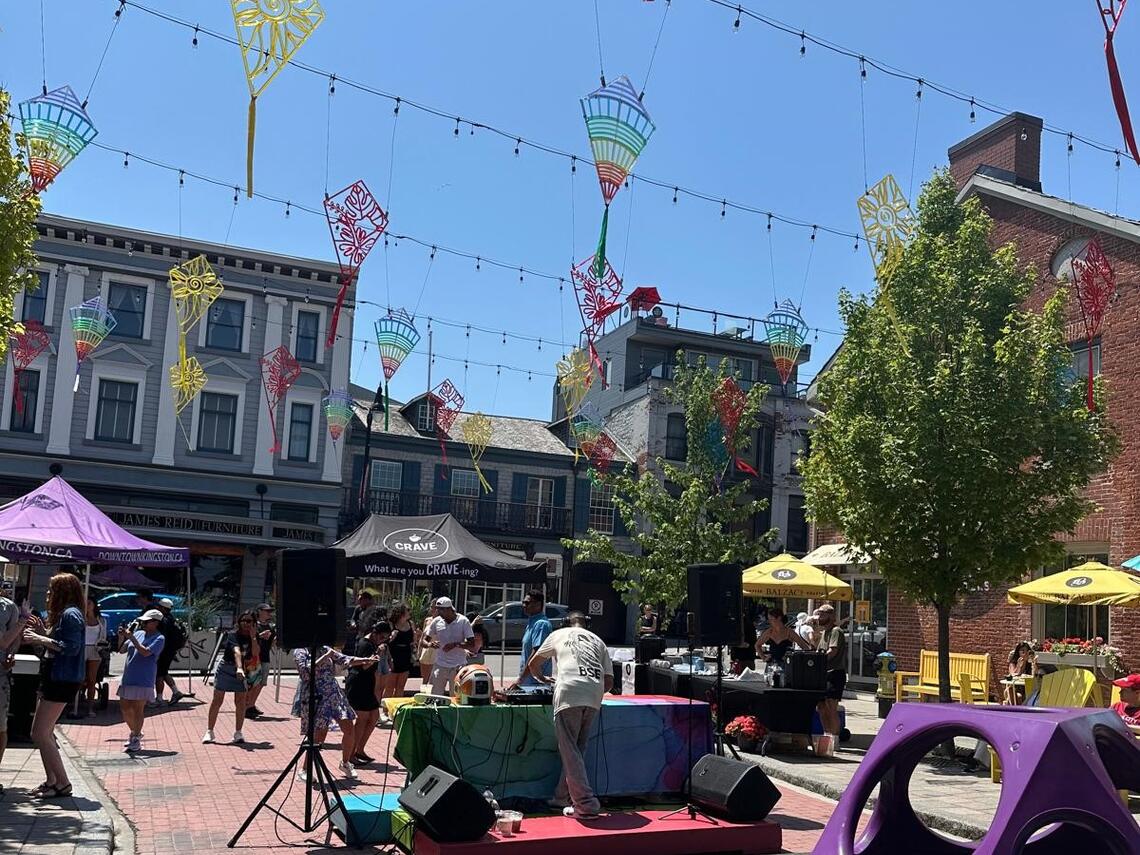 An outdoor summer event in downtown Kingston with colorful kite-shaped decorations hanging overhead, people gathered around a small stage with a DJ.  