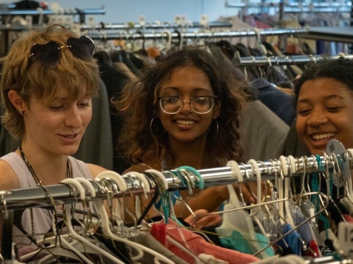 Three university-aged students smiling as they browse racks of gently used clothes URL: https://today.wayne.edu/news/2022/10/26/the-w-pantry-and-thrift-shop-working-to-end-student-clothing-and-food-insecurity-49435
