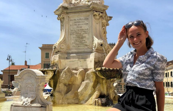 A woman sitting on a railing smiling. Behind her is a decorative fountain.
