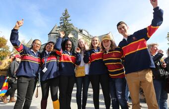 Seven Queen’s University students in striped rugby shirts cheer with raised arms at a sunny campus celebration in front of a stone building.
