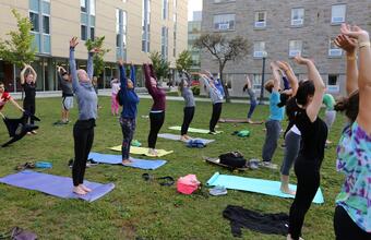 Students participate in an outdoor group yoga class on a grassy lawn between campus buildings, standing on mats with their arms raised overhead.