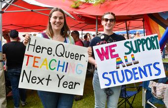 Two people stand at a Pride event holding handmade signs.