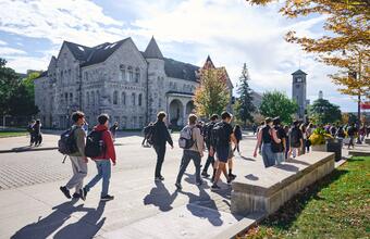 Students with backpacks walk in front of Ontario Hall at Queen's university surrounded by trees with early autumn leaves.