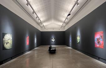 Wide view of a dark-walled gallery with colourful sci-fi collages, a central bench, polished concrete floor, and vaulted ceiling with track lighting.