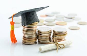 Stacked coins arranged in ascending height with a graduation cap on the tallest stack and a rolled diploma tied with string in the foreground, symbolizing the financial cost or investment of higher education.