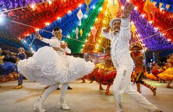 Dancers in colorful costumes perform under a rainbow canopy of paper decorations and lights at a festive Brazilian June festival celebration. 