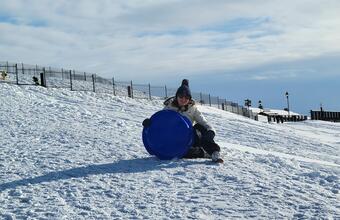 Person in winter gear sitting on a snowy hill, smiling and holding a blue sled, with a fence and cloudy sky behind.