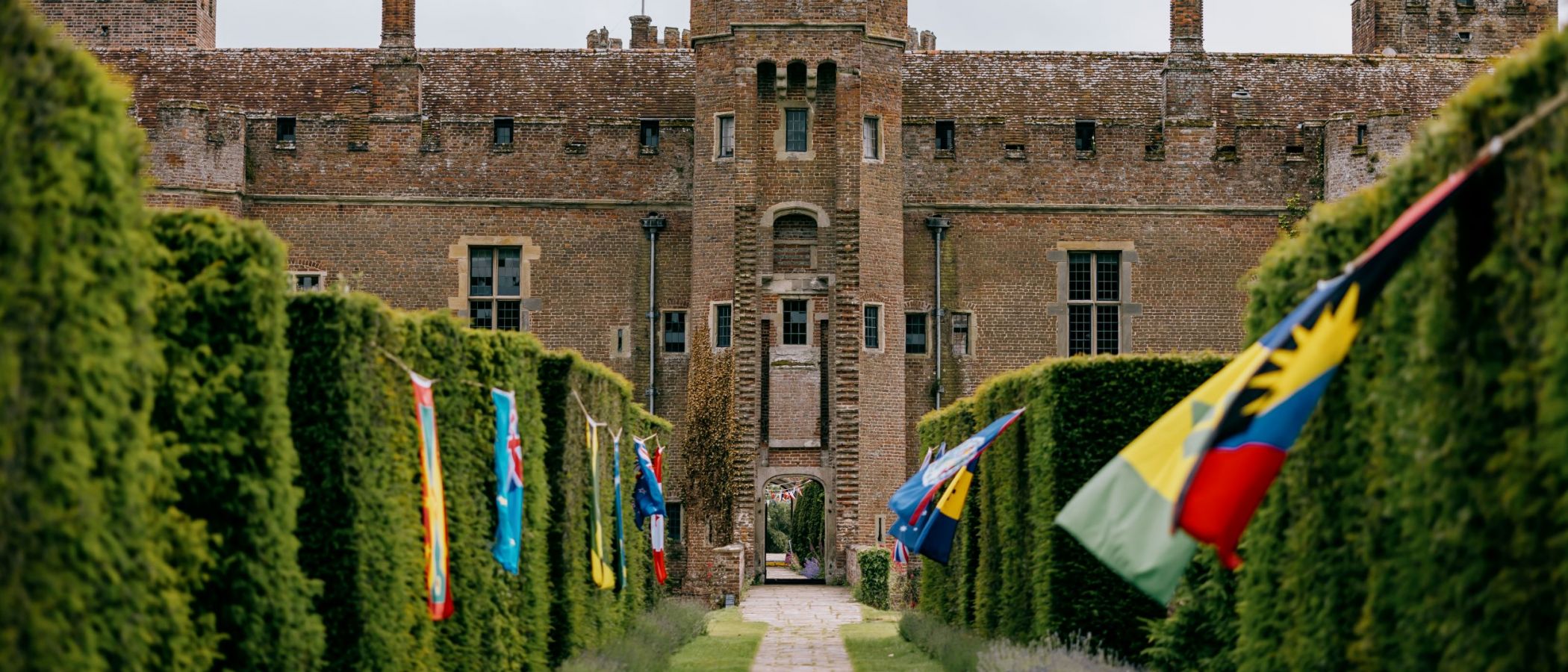 An image of Herstmonceux Castle, UK with various flags down the sides of the hedged walkway 