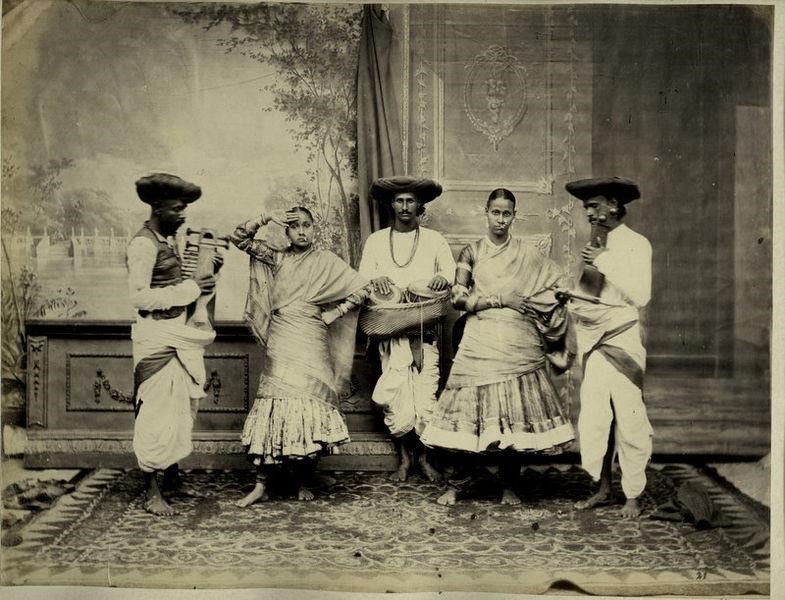 A sepia-toned image of three men and two women. The women are in ornate dress. 