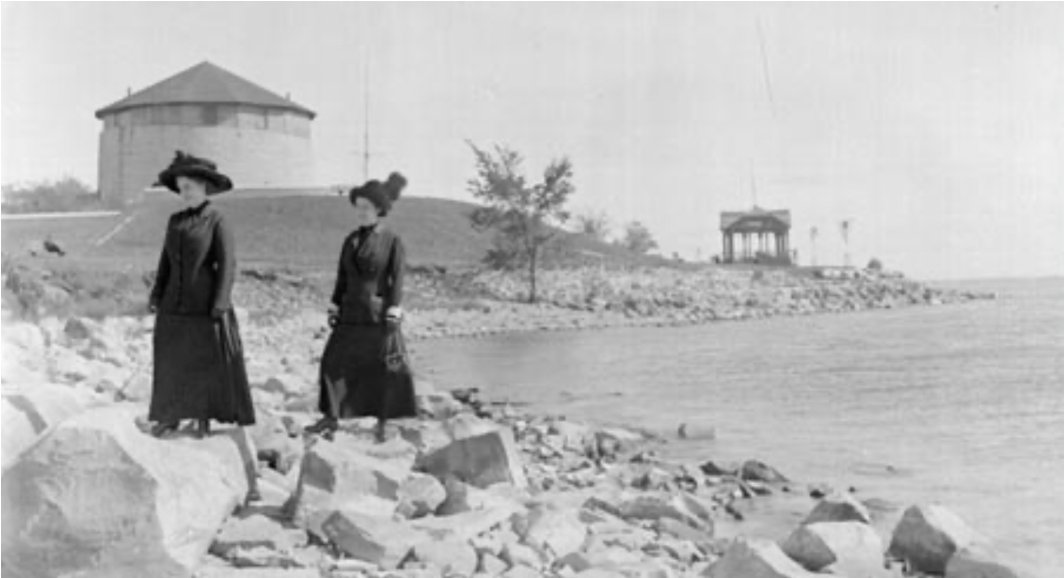 A black and white photograph of two women standing on the rocks at Macdonald Park on the shore of Lake Ontario with Murney Tower in the background.