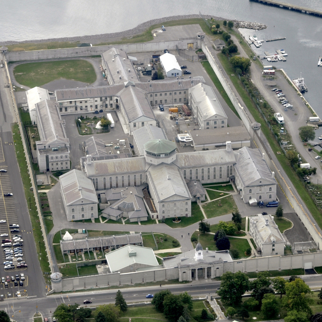 An aerial image of Kingston Penitentiary where you can see the stone entryway, two central "t" shaped buildings, and a number of various outbuildings with a parking lot on the left 