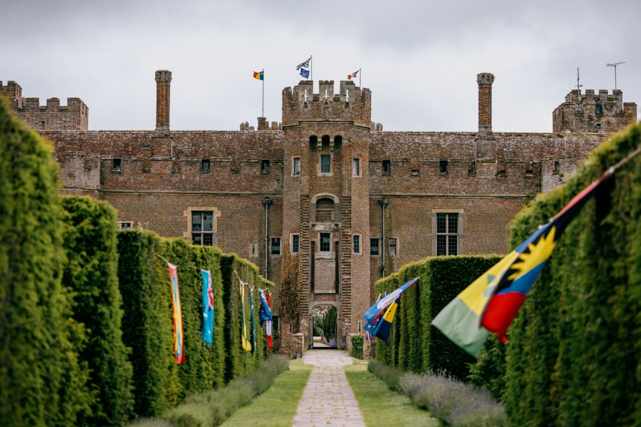 An image of Bader College featuring a stone walkway leading to the red brick castle and red, yellow, and blue flags on the hedges that line the walkway