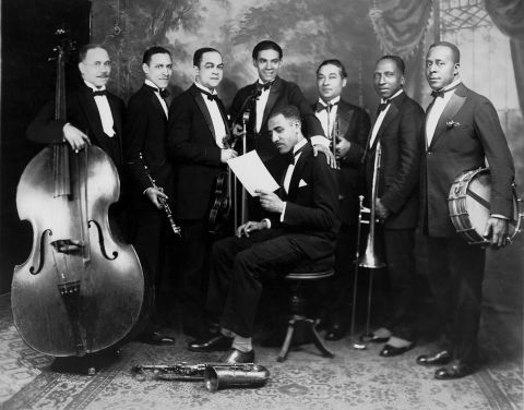 An image of Lou Hooper reading music with seven men surrounding him holding instruments