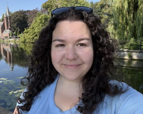 Kaitlyn is pictured smiling in front of a river in Bruges, Belgium, a small portion of a dutch style building can be seen on the left side of the image. She is white with freckles on her nose and cheeks. Her hair is long, dark, and curly. She wears a blue t-shirt and sunglasses on the top of her head.