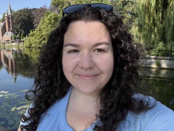 Kaitlyn is pictured smiling in front of a river in Bruges, Belgium, a small portion of a dutch style building can be seen on the left side of the image. She is white with freckles on her nose and cheeks. Her hair is long, dark, and curly. She wears a blue t-shirt and sunglasses on the top of her head.