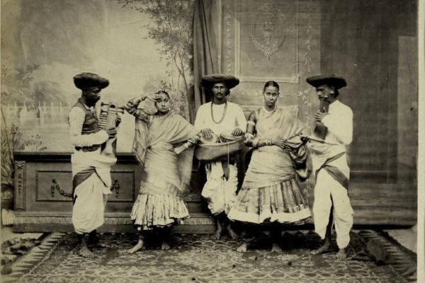 A sepia-toned image of three men and two women. The women are in ornate dress. 