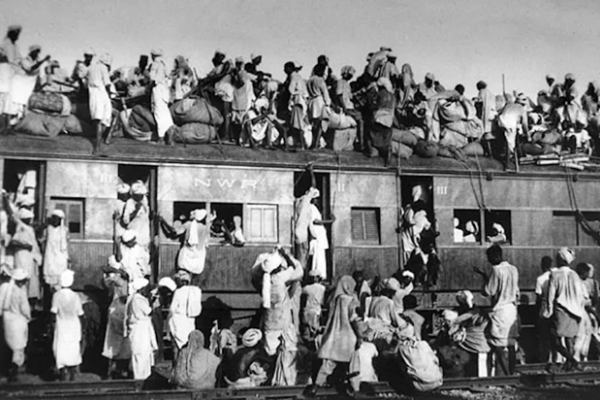 A black and white image of a full train with people standing on the roof and hanging off the sides of the cars