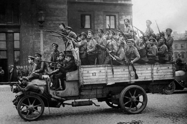An image of a group of men holding guns on an old truck riding through a city square