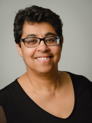 Photo of Dr. Imaan Bayoumi who is smiling. She is wearing black frame glasses with a white shirt and has short black hair 