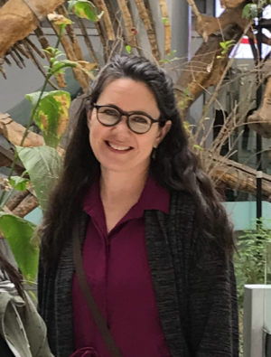 Photo of Dr. Colleen Davison who is smiling and standing in front of some plants. She is wearing black frame glasses with a purple shirt and grey cardigan and has long brown hair
