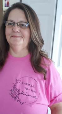 Photo of Michele Cole who is smiling with her mouthed closed. She is wearing glasses with a pink shirt that reads, "Choose to be Kind", and has long brown hair