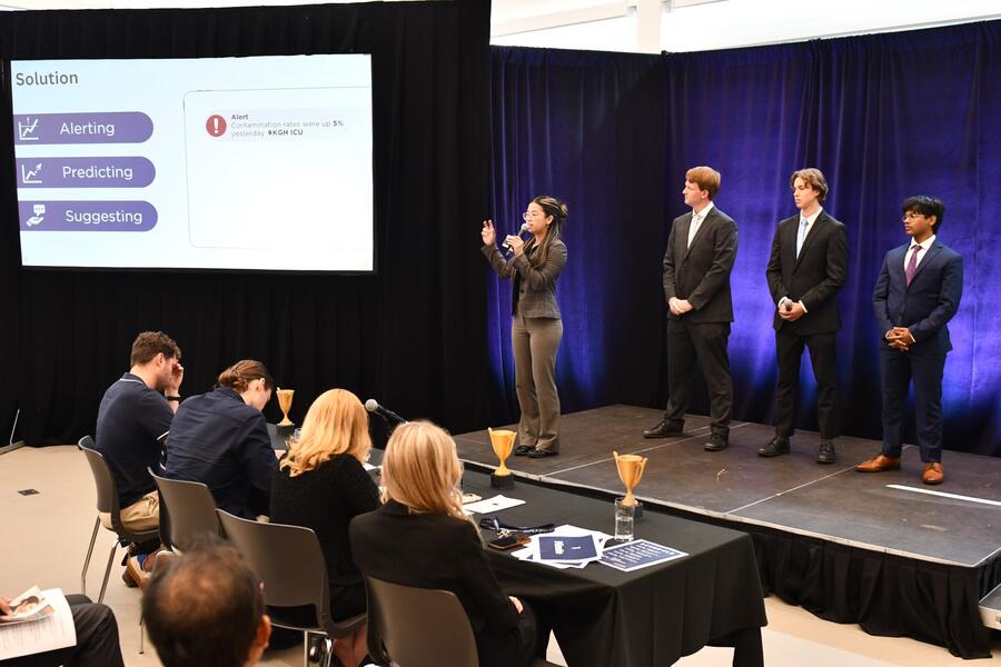 A team of four young professionals in business attire presents on stage during a pitch competition. One woman is speaking into a microphone and gesturing with her hands, while three men stand beside her attentively. Behind them, a slide is projected with the title “Solution” and three points: “Alerting,” “Predicting,” and “Suggesting.” Judges sit at a table in front of the stage, listening and taking notes, with small gold trophies placed on the table and stage.