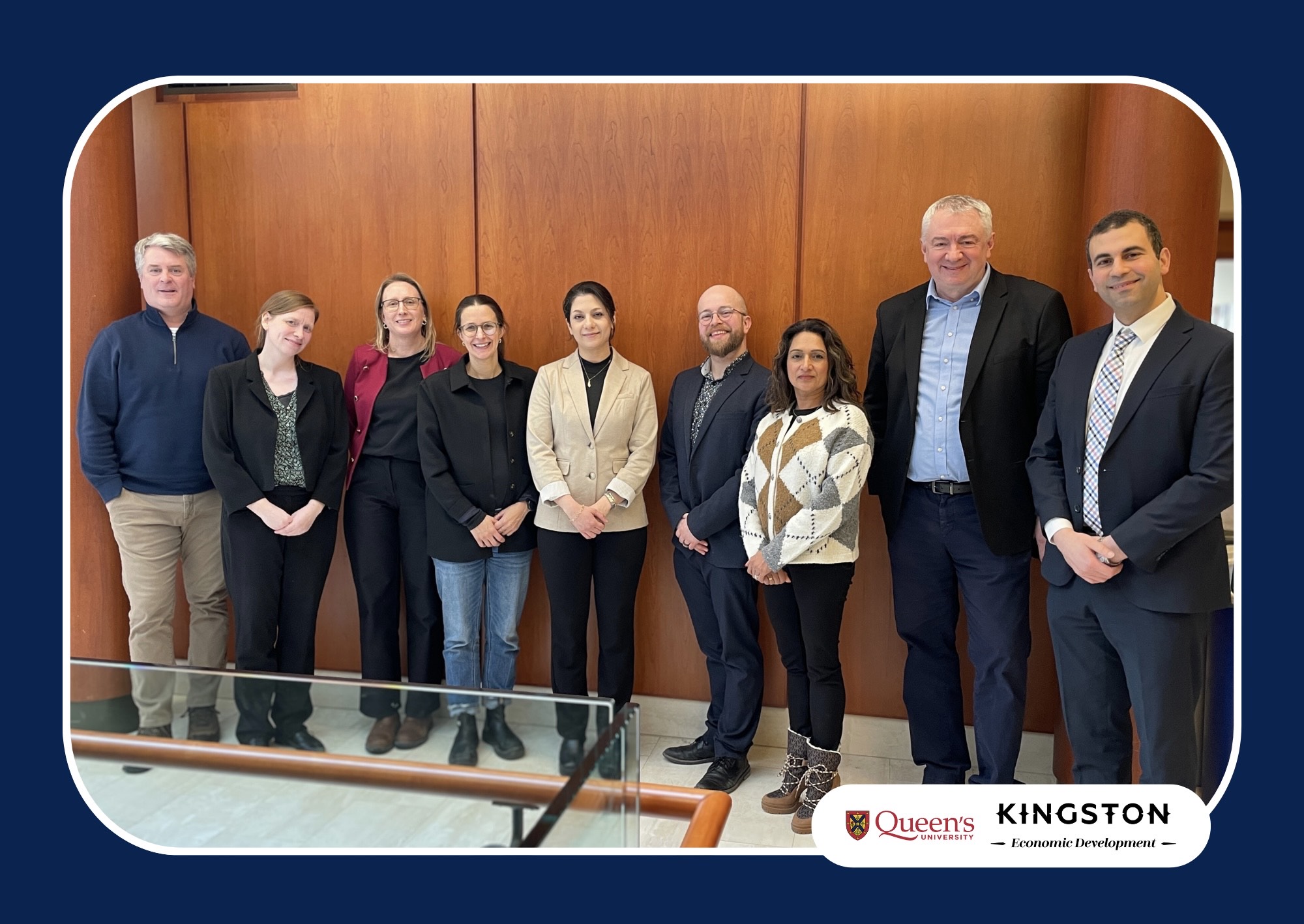 A group of nine people standing in a row inside a wood‑paneled room, posing for a group photo. A railing is visible in the foreground, and Queen’s University and Kingston Economic Development logos appear in the bottom right corner