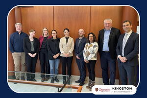 A group of nine people standing in a row inside a wood‑paneled room, posing for a group photo. A railing is visible in the foreground, and Queen’s University and Kingston Economic Development logos appear in the bottom right corner