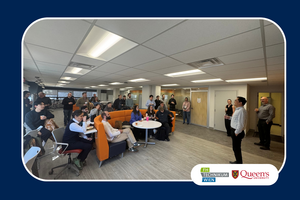 A group of people gather in a bright office space with modern furniture, listening to a speaker. The atmosphere is engaged and informal. Logos for FH Technikum Wien and Queen's University are visible.