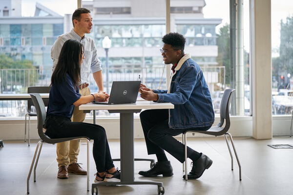 Three colleagues are gathered at a table. Two are seated with a laptop and one stands, engaged in discussion. Large windows reveal an urban backdrop.