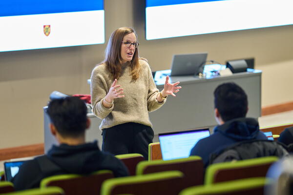 A woman in a beige sweater passionately speaks to students in a lecture hall. She's gesturing with her hands, while students with laptops listen attentively.