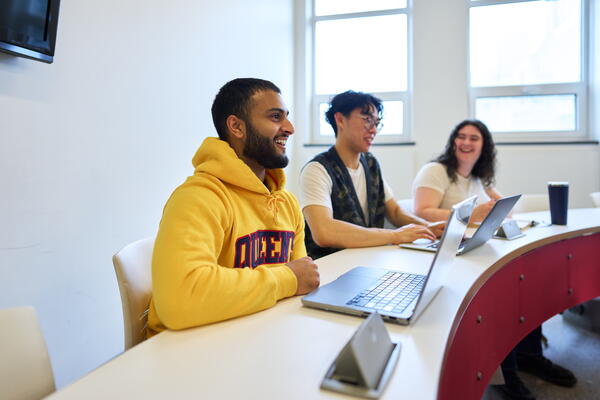 Three people sit at a curved table with laptops, smiling and engaging in conversation. The room is bright, suggesting a positive, collaborative atmosphere.