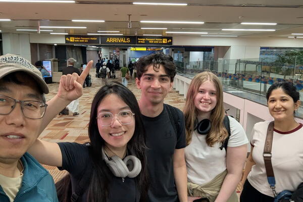 A group of five people smiling at an airport's arrival area. One person points to the "Arrivals" sign. The atmosphere is cheerful and welcoming.