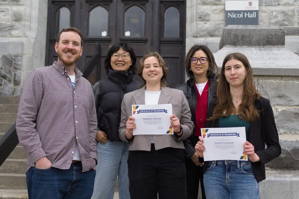 A group of five smiling people stand on stairs outside a building named Nicol Hall. Two individuals hold certificates of recognition. The mood is celebratory.