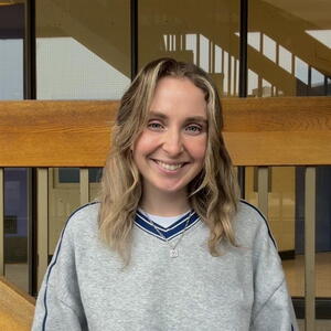 Madie with wavy hair wearing a gray sweatshirt stands in front of a wooden structure. Warm lighting creates a welcoming atmosphere.