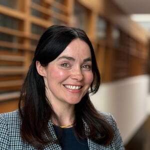 Rachel poses in checked blazer smiling, standing in a modern hallway with wooden paneling and natural light.